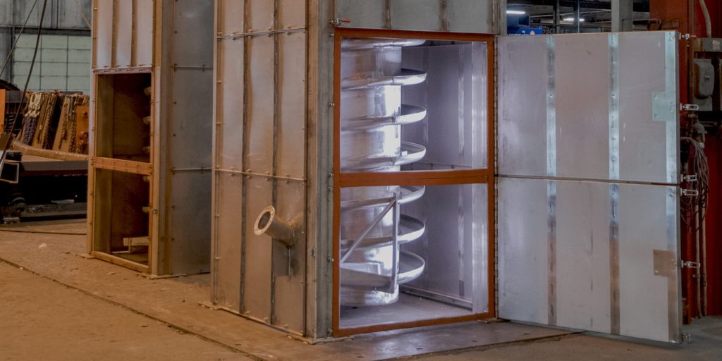 Industrial machinery with a large metallic chamber and exposed spiral blades inside, set in a factory floor scene.