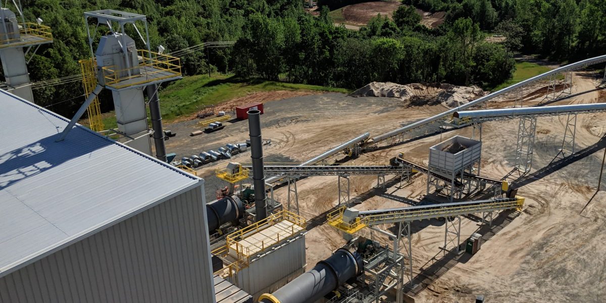 Aerial view of an industrial processing site with conveyors, metal towers, and yellow safety rails on a dusty yard surrounded by trees.