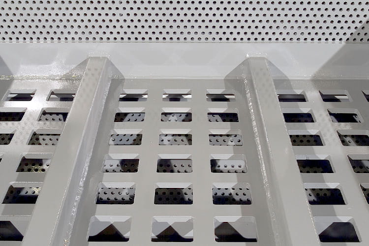Industrial metal perforated floor/grating with square holes and two supporting beams, viewed from above, in a workshop setting.
