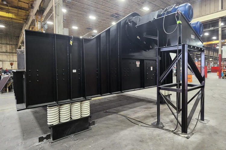 Industrial factory setting with a long black conveyor system supported by spiral-spring legs, blue motors on top, and cables on the floor.